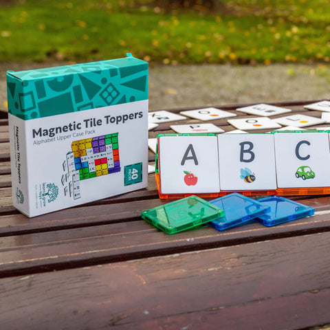 A set of Learn & Grow Rainbow Magnetic Tiles Toppers - Alphabet Upper Case Pack 40pc is displayed on a wooden bench, with three tiles in the forefront showing the letters A, B, and C, each paired with corresponding images of an apple, a bee, and a car. Perfect for early literacy instruction, the background features a grassy area.