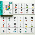 A set of 40 magnetic tile toppers with uppercase letters and illustrations from the Learn & Grow Rainbow Magnetic Tiles Toppers - Alphabet Upper Case Pack are arranged neatly. Letters A to Z appear on individual white tiles, each accompanied by a corresponding animal or object, making them ideal for early literacy instruction. The box for the Alphabet Upper Case set from Learn & Grow is visible in the top left corner.