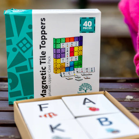 A box of "Learn & Grow Rainbow Magnetic Tiles Toppers - Alphabet Upper Case Pack 40pc" from Learn & Grow, featuring a colorful illustration of upper-case alphabet tiles, is displayed on a wooden surface. In front of the box, an open package reveals some of the magnetic letter tiles, ideal for early literacy instruction. The box indicates it contains 40 pieces.