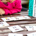 A young child in a pink sweater is playing with alphabet cards from the Learn & Grow Rainbow Magnetic Tiles Toppers - Alphabet Upper Case Pack on a wooden table. The cards, part of an early literacy instruction set, display letters like D, A, N, H, and M with corresponding images. A box labeled "Learn & Grow" is partially visible on the table.