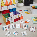 A vibrant educational display features the Learn & Grow Rainbow Magnetic Tiles Toppers - Alphabet Upper Case Pack 40pc, forming structures that spell out "RAIN" and "HELLO," alongside animal cards. Various Alphabet Upper Case and picture cards, including vowels, are scattered on the carpeted floor along with other educational toys for early literacy instruction.