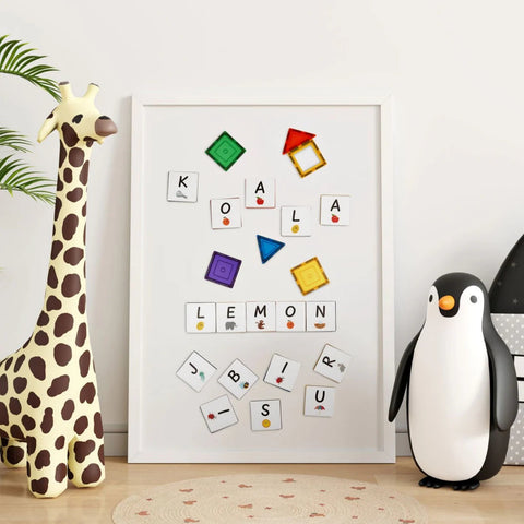 A children's room features a whiteboard adorned with Learn & Grow Rainbow Magnetic Tiles Toppers from the Alphabet Upper Case Pack, forming words like "KOALA" and "LEMON" to aid early literacy. A plush giraffe stands on the left, while a penguin-shaped storage bin sits on the right. An Alphabet Upper Case dot-patterned rug covers the floor.