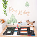 A children's room decorated with a leafy green plant hanging from the top left features wooden words "Play all day" on the wall with tree decals and a stream. Below is a play table, perfect for imaginative play, showcasing a toy car on a Learn & Grow Rainbow Magnetic Tiles Toppers - Road Pack 40pc track alongside.