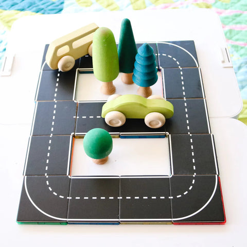 The Learn & Grow Rainbow Magnetic Tiles Toppers - Road Pack 40pc is used to arrange an imaginative play roadway scene. This setup features a yellow toy bus, a green toy car, and three green trees in different shapes—one conical and two spherical. The backdrop includes part of a quilted blanket, creating endless adventures with this Road Pack from Learn & Grow.