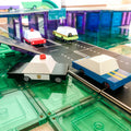 Toy cars, including a police car, are on a play road made of colorful Learn & Grow Rainbow Magnetic Tile Toppers - Road Pack 40pc. The setup features a small bridge, various road signs, and a vibrant backdrop resembling a miniature cityscape—perfect for imaginative play with the Road Pack from Learn & Grow.