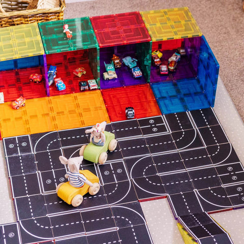 A children's play area with colorful, translucent building blocks forming cubbyholes that feature Learn & Grow Rainbow Magnetic Tiles Toppers. Toy cars are placed inside the cubbyholes, and the floor is covered with a Learn & Grow Road Pack 40pc featuring various tracks. Two animal figurines on toy scooters enjoy imaginative play on the road map.