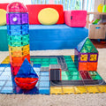 A vibrant play area showcases the Learn & Grow Rainbow Magnetic Tile Toppers - Road Pack 40pc intricately arranged to create a towering clock, a sailboat with triangular sails, a red and yellow house, a cube, and an interconnected road on a mat. In the background, there's a blue couch adorned with yellow and red cushions, providing an ideal setting for imaginative play.