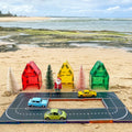 A beach scene sets the stage for imaginative play with the Learn & Grow Rainbow Magnetic Tiles Toppers - Road Pack (40 pieces). This setup includes a miniature toy town with colorful toy houses, tiny trees, and a Santa figurine. The road pack features three magnetic toy cars in blue, yellow, and green navigating curved roads. In the background, the ocean and sandy shore provide the perfect backdrop for creative adventures.