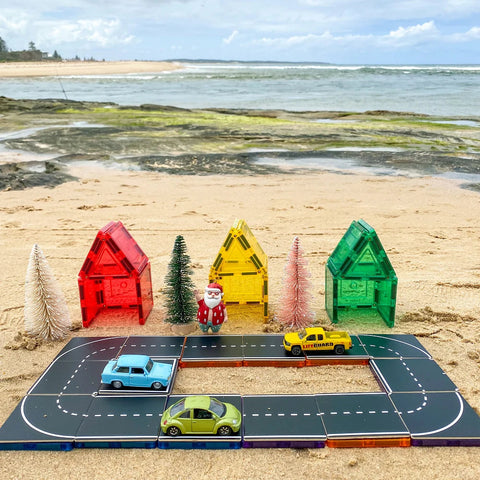 A beach scene sets the stage for imaginative play with the Learn & Grow Rainbow Magnetic Tiles Toppers - Road Pack (40 pieces). This setup includes a miniature toy town with colorful toy houses, tiny trees, and a Santa figurine. The road pack features three magnetic toy cars in blue, yellow, and green navigating curved roads. In the background, the ocean and sandy shore provide the perfect backdrop for creative adventures.