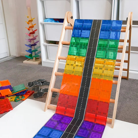 The Learn & Grow wooden play structure features a ramp adorned with an array of colorful, translucent tiles from the Learn & Grow Rainbow Magnetic Tiles Toppers - Road Pack 40pc, creating a vibrant rainbow pathway. This imaginative roadway descends elegantly from the structure, ideal for creative play. In the background, additional magnetic tiles and a vibrant toy tree can be seen, adding to the playful scene.