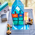 A vibrant magnetic tile creation, made using the Learn & Grow Rainbow Magnetic Tiles Toppers - Road Pack 40pc, forms a house on a black grid mat, perfect for imaginative play. Nearby, wooden toy carriages are transporting peg people. On the left side of the mat, there's a box labeled "Magnetic Tile Toppers" and a stack of cards from the Road Pack.