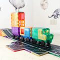 A Learn & Grow Rainbow Magnetic Tiles Toppers - Road Pack 40pc toy truck and trailer built from vibrant, translucent blocks are set on a black playmat designed to resemble a road. The background wall is adorned with animal decals, including a raccoon and a deer, creating an ideal environment for imaginative play. An electrical outlet is also visible on the wall.