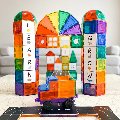 Using the Learn & Grow Rainbow Magnetic Tiles Toppers from the Road Pack (40 pieces), a vibrant archway with two towers has been constructed. The left tower proudly displays "LEARN" while the right tower showcases "GROW." Positioned in front of this imaginative setup is a blue and orange toy truck, sparking creativity against a softly blurred living room background.