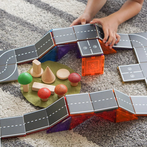 A child's hands are assembling a miniature road set with various geometric blocks, Learn & Grow Rainbow Magnetic Tiles Toppers - Road Pack 40pc on a carpeted floor. There are toy trees and wooden shapes arranged in the center, depicting a small park or play area perfect for imaginative play.