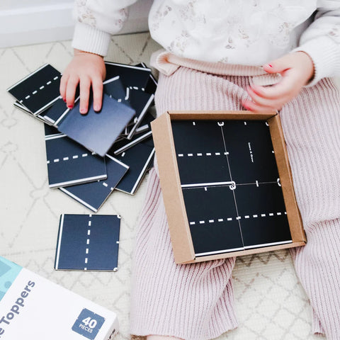 A child sits on a cream-colored carpet, engaged in imaginative play with black square tiles featuring white road markings from the Learn & Grow Rainbow Magnetic Tiles Toppers - Road Pack 40pc. The child, with pink nail polish, wears a white sweater and light pink pants. Next to them is an open cardboard box filled with more tiles from the Learn & Grow Road Pack.