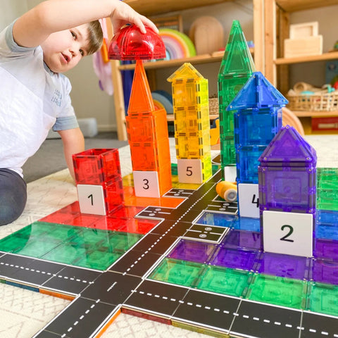 A child is engrossed in imaginative play, creating colorful, translucent structures with Learn & Grow Rainbow Magnetic Tiles numbered 1 to 5 on a Road Pack playmat that features a detailed road pattern. Reaching for a red dome from the Learn & Grow Rainbow Magnetic Tiles Toppers set, the child carefully places it atop an orange structure. The background shows various toys and shelves.