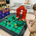 A child engaged in imaginative play arranges Learn & Grow Rainbow Magnetic Tiles Toppers - Road Pack 40pc and small toy cars on a wooden table. The tiles form structures like a red archway and green formations. Shelves of more toys are visible in the background, with Learn & Grow's Magnetic Tile Toppers adding extra fun to their Road Pack adventures.