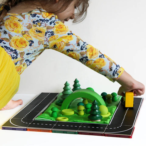 A child dressed in a yellow floral top and yellow skirt engages in imaginative play with the Learn & Grow Rainbow Magnetic Tiles Toppers - Road Pack 40pc, featuring a black road with white lines, green toy trees, and hills. The child pushes a small yellow vehicle along the Road Pack, enriched with Magnetic Tile Toppers from Learn & Grow for added fun.