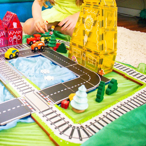 A child playing with the 36-piece Learn & Grow Rainbow Magnetic Tiles Toppers - Train Pack on a play mat. The scene includes a roadway, small buildings, and train tracks. Toy cars and a green tree are also visible. The child's upper body and hands are seen in the background as they interact with the set from Learn & Grow.