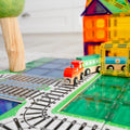 A brightly colored toy train from the Learn & Grow Rainbow Magnetic Tiles Toppers - Train Pack 36pc travels on a track laid out on a play mat. Surrounding the train are multicolored magnetic tiles from the set, arranged to form structures like a house and a tree, simulating a playful, miniature landscape complete with Magnetic Tile Toppers.