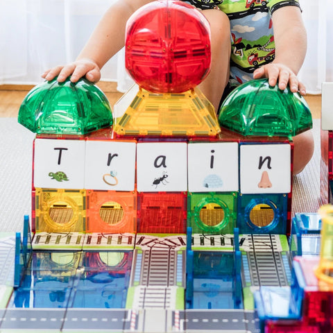 A child in vibrant pajamas enjoys playing with the Learn & Grow Rainbow Magnetic Tiles Toppers - Train Pack 36pc, assembling magnetic tiles into a structure that displays the word "train." This 36-piece set from Learn & Grow features both transparent and colorful tiles, each illustrated with objects corresponding to each letter (turtle, ring, ant, ice cream, nose).