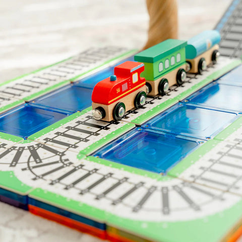 A wooden toy train from the Learn & Grow Rainbow Magnetic Tiles Toppers - Train Pack 36pc, which includes a red engine, green carriage, and blue caboose, travels on a track with blue tiles underneath. The track is set on a green and white base with various curves and straight segments as part of this intricate 36 piece set by Learn & Grow.