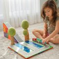 A young girl with long brown hair sits on a patterned rug, engrossed in play with her 36-piece train set. The colorful tracks, complete with a ramp and two tree figurines, are made even more exciting thanks to the Learn & Grow Rainbow Magnetic Tiles Toppers - Train Pack 36pc. Sheer white curtains and a soft, neutral-toned room form the serene backdrop.