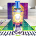 The Learn & Grow Rainbow Magnetic Tiles Toppers - Train Pack 36pc features a colorful train made of translucent, multicolored plastic blocks illuminated by a bright light at the front. It's placed on a patterned track composed of similar tiles from the 36-piece set. The background includes out-of-focus objects and a neutral setting, highlighting the vibrant scene.