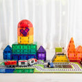 The Learn & Grow Rainbow Magnetic Tiles Toppers - Train Pack (36 pieces) by Learn & Grow features colorful magnetic tiles that can be assembled into geometric structures resembling buildings, such as a rocket-like tower and pyramids. A 36-piece toy train pack runs on a track around the buildings with two wooden figurines standing nearby, all set against a backdrop of white curtains.