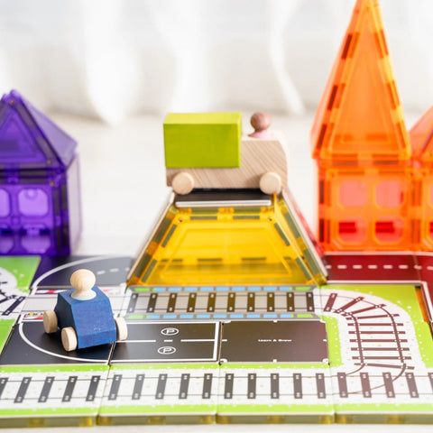 A vibrant scene of children's toys includes a wooden toy car with a peg doll driver, the Learn & Grow Rainbow Magnetic Tiles Toppers - Train Pack 36pc featuring pieces in orange, purple, and yellow, and a toy truck carrying blocks. The tile mat features roads and train tracks.