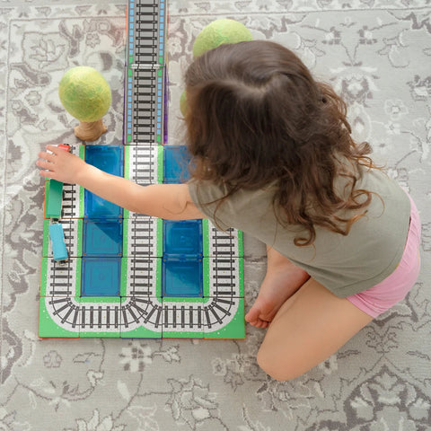 A young child with curly hair, wearing a gray shirt and pink shorts, is playing with the Learn & Grow Rainbow Magnetic Tiles Toppers - Train Pack 36pc on a patterned rug. The set includes track pieces, blue blocks, green balls, and a miniature train. The child is reaching for one of the blue blocks.