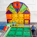 A colorful magnetic tile structure resembling a house stands on a mat. In front of the structure, a toy train from the Learn & Grow Rainbow Magnetic Tiles Toppers - Train Pack 36pc and a wooden figure in a car are positioned on tracks and roads. The Learn & Grow set features vibrant tiles in yellow, orange, blue, and green.