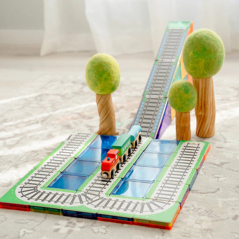 A Learn & Grow Rainbow Magnetic Tiles Toppers - Train Pack 36pc set is displayed on a light-colored carpet, showcasing a red and green engine, a green cargo car, and an extensive track that includes an elevated bridge with ascending and descending paths. The set also features two unique toy trees with spiral trunks and green canopies positioned alongside the track.