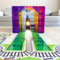 The Learn & Grow Rainbow Magnetic Tiles Toppers - Train Pack 36pc forms a vibrant tunnel over the train track, brightly illuminated from the inside. The tracks are laid out on a play mat, with a grey sofa in the background. This product from Learn & Grow adds an extra layer of fun to playtime!