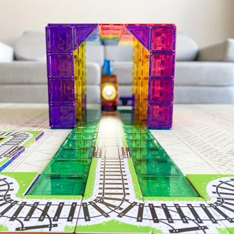 The Learn & Grow Rainbow Magnetic Tiles Toppers - Train Pack 36pc forms a vibrant tunnel over the train track, brightly illuminated from the inside. The tracks are laid out on a play mat, with a grey sofa in the background. This product from Learn & Grow adds an extra layer of fun to playtime!