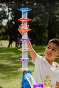 A young boy outdoors, reaching up to touch the top of a tall tower he built using the colorful translucent pieces from the Magblox Magbrix Rainbow Ball Run 98pc set by Magblox. The tower features blue, orange, and clear segments. Trees and greenery can be seen in the background. The boy is wearing a white T-shirt and smiling, showcasing STEAM learning in action.