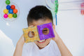 A young child smiles, holding two colorful translucent pieces from the Magblox Magbrix Rainbow Ball Run 98pc set over their eyes. Brightly colored balls are visible in the background on the left, and a clear plastic play tunnel extends from the right side. The scene is brightly lit and playful, perfect for fostering fine motor skills with this engaging product from Magblox.