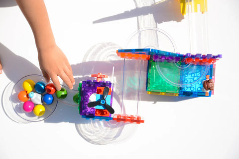 A child's hand reaches for colorful plastic balls in a bowl next to the Magblox Magbrix Rainbow Ball Run 98pc construction set by Magblox, which is made of transparent and brightly-colored interlocking pieces featuring a fan and various pathways. This engaging activity promotes fine motor skills and STEAM learning on a white surface.