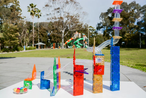 The Magblox Magbrix Rainbow Ball Run 98pc set is a colorful and engaging marble run, comprised of tall, transparent structures interconnected by tracks. Set up on a white surface outdoors amidst grass, trees, a playground, and clear skies, it offers an inviting backdrop. Four marbles are neatly gathered on the left side, encouraging fine motor skills development and STEAM learning.

