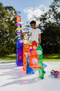 A child plays outside with colorful magnetic building tiles from the Magblox Magbrix Rainbow Ball Run 98pc set by Magblox, creating tall, intricate structures. The tiles are a mix of vibrant colors, including red, green, blue, and orange. The child, wearing a white shirt and focused on STEAM learning and fine motor skills, adds more pieces to the arrangement.