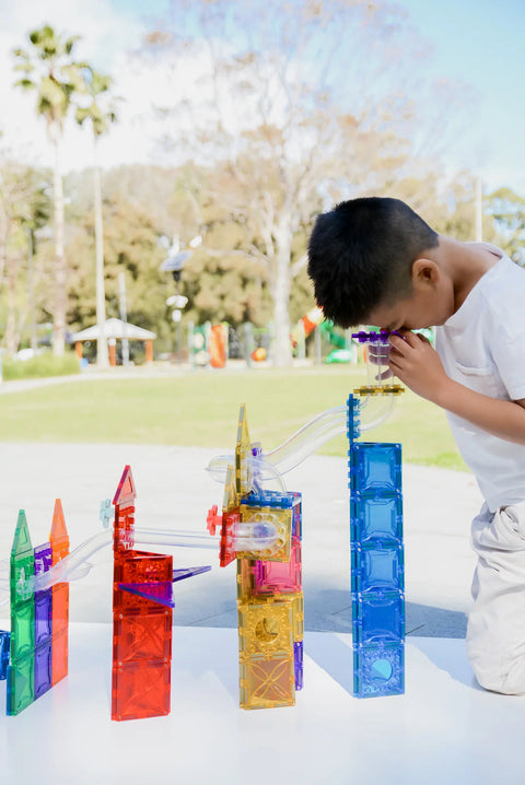 A child kneels on the ground, focusing intently on a vibrant creation of Magblox Magbrix Rainbow Ball Run 98pc tiles forming towers of various shapes and heights. This engaging activity encourages STEAM learning and fine motor skills, all set against a sunny park backdrop with trees and a walkway.