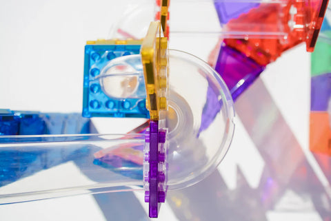 A close-up of the Magblox Magbrix Rainbow Ball Run 98pc by Magblox displays its vibrant, transparent plastic tubes and a variety of bright interlocking pieces in yellow, purple, red, and blue. Set up on a white surface, the configuration showcases shadows cast by the pieces, promoting STEAM learning while enhancing fine motor skills.