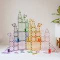 A vibrant geometric structure built from transparent magnetic tiles in a variety of shapes and pastel hues, including purple, green, orange, yellow, and blue. This colorful creation beautifully showcases the Magblox Pastel Pack 66pc from Magblox against a white background, with a plant in a white pot positioned on the right.