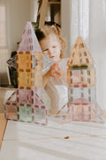 A young child with brown hair, wearing a white polka-dot dress, stands at a table, looking inquisitively at two colorful, translucent plastic block towers built from the Magblox Pastel Pack 66pc. Sunlight streams through a window, illuminating the room and enhancing their STEAM learning experience.