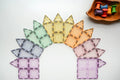 A symmetrical arrangement of colorful, transparent **Magblox** tiles from the **Magblox Pastel Pack 66pc** is displayed on a white surface, forming a crescent shape. In the top-right corner, there is a wooden tray containing variously colored spools, promoting eco-friendly STEAM learning.