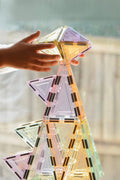 A child's hands are seen placing a colorful, geometric building piece from the Magblox Pastel Pack 66pc by Magblox onto a tall, intricate tower made of translucent magnetic tiles in various shapes and colors. The background shows a blurred wooden fence and window, emphasizing the focus on this eco-friendly STEAM learning toy.