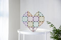 A heart-shaped structure made of colorful translucent geometric tiles, from the eco-friendly Magblox Pastel Pack 66pc by Magblox, stands on a small, round, white table. The background features a white curtain and a potted green plant, adding a serene ambiance to this STEAM learning scene.