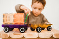 A young child is deeply engaged in creative play with the colorful Magblox Twin Car Set on a wooden table. The translucent toy trucks, featuring purple and orange hues and magnetic car bases from Magblox, captivate their focus. The plain white background highlights the child's fascination with the toys.