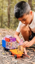 A young boy with short dark hair is kneeling on the ground in a forested area, intently engaged in creative play with colorful translucent magnetic building blocks from the Magblox Twin Car Set by Magblox, assembling them on magnetic car bases. He wears a white t-shirt and dark shorts, surrounded by fallen leaves.