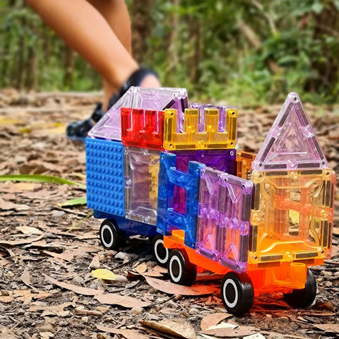 A toy train made from vibrant, see-through Magblox Twin Car Set building blocks rests on a forest floor scattered with dry leaves and twigs. In the background, an out-of-focus child in blue shoes is walking. The magnetic car bases from Magblox spark hours of imaginative play.